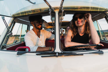 Cheerful man and woman smiling and sitting inside vintage van while traveling in countryside on sunny day together - ADSF06297