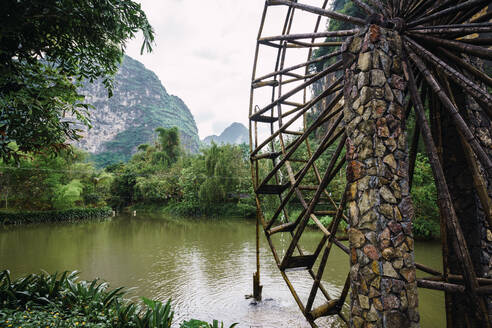 Schönes hölzernes Wasserrad einer alten steinernen Wassermühle, umgeben von Bäumen am Ufer des Quây Son Flusses mit bewölktem Himmel und herrlichen Bergen im Hintergrund in der chinesischen Provinz Guangxi - ADSF06330