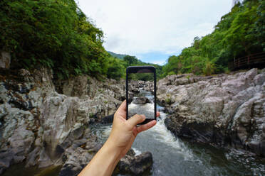 Crop-Tourist mit Smartphone und Aufnahme eines Wasserlaufs zwischen Felsen im Yanoda-Regenwald, China - ADSF06333