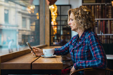 Side view of woman in plaid T-shirt sitting in cafe with cup of coffee on table and using mobile phone - ADSF06341
