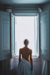 Rückenansicht einer nackten jungen Frau mit weißem Handtuch, die nach dem Duschen am geöffneten Fenster steht - ADSF06342