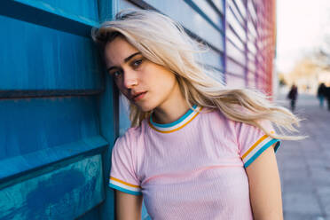 Junge blonde Frau steht auf der Straße und schaut in die Kamera, während sie sich an eine blaue Wand lehnt. - ADSF06345