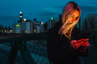 Hübsche blonde Frau, die in der Abenddämmerung auf der Straße steht und ihr Smartphone benutzt. - ADSF06351