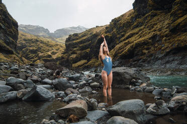 Young beautiful female standing in swimming suit in mountain river with big rocks with hands up - ADSF06352