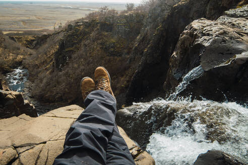 Crop Beine der männlichen sitzen auf dem Gipfel des Berges in der Nähe von Wasserfall in Winterkleidung - ADSF06370