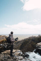 Rückenansicht eines männlichen Fotografen in Winterkleidung mit Rucksack und Kamera, der den Meerblick vom Berg aus genießt - ADSF06371