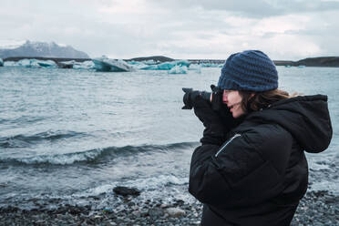Seitenansicht einer Frau in Freizeitkleidung, die mit einer Kamera eine wunderschöne Eismeerlandschaft in Island fotografiert. - ADSF06374