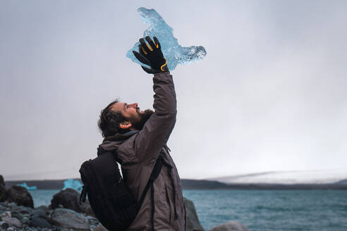 Seitenansicht eines glücklichen, bärtigen Mannes mit Rucksack, der einen Eisbrocken über dem Kopf hält, in der Landschaft Islands. - ADSF06375