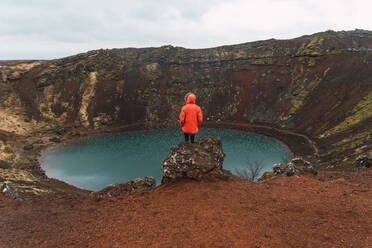 Back view of tourist in red coat standing on rock against small blue lake in basin of mountain in Iceland. - ADSF06378