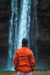 Rückenansicht eines Mannes in orangefarbenem Wintermantel, der mit den Händen in den Taschen auf einen Wasserfall blickt, Island. - ADSF06380
