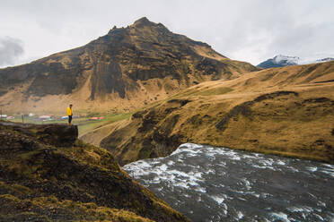 Blick auf einen Touristen, der auf einem Felsen über fließendem Wasser steht, mit malerischen Bergen im Hintergrund, Island. - ADSF06392