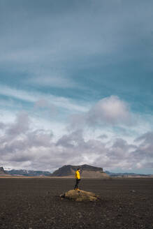 Anonymer Mann steht auf einem Felsen in einer felsigen Ebene mit Bergen im Hintergrund unter bewölktem Himmel, Island. - ADSF06394