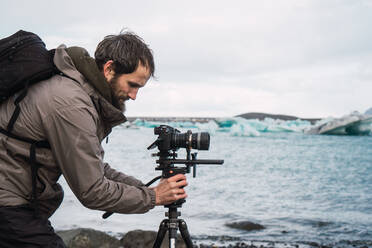 Seitenansicht eines Mannes in Outdoor-Kleidung mit Rucksack, der eine Fotokamera auf ein Stativ stellt, um eine schöne kalte Meereslandschaft zu fotografieren. - ADSF06408