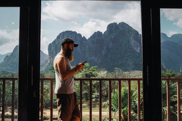 Seitenansicht eines bärtigen Mannes mit Smartphone auf einem Balkon vor dem Hintergrund der Berge. - ADSF06439