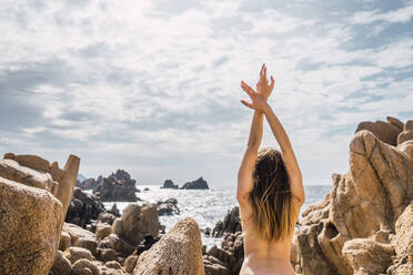 Rückenansicht einer nackten Frau mit erhobenen Händen, die sich an den Küstenfelsen am Ozean streckt. - ADSF06449