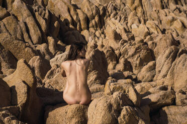 Rückenansicht einer nackten Frau, die auf einem Felsen sitzt und mit den Händen hinter dem Rücken posiert. - ADSF06450