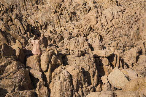 Rückenansicht einer nackten Frau, die auf einem Felsen sitzt und mit den Händen hinter dem Rücken posiert. - ADSF06451