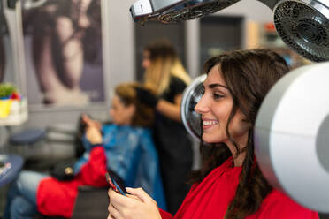 Side view of cheerful lady holding mobile phone and drying hairs in hairdressing salon - ADSF06459