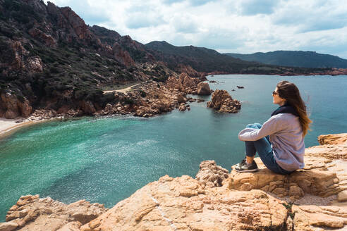 Seitenansicht einer Touristin, die auf einem Felsen sitzt und auf eine Bucht und einen Hügel blickt. - ADSF06493