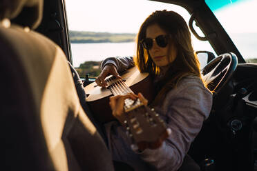 Attraktive junge Frau spielt Gitarre und sitzt im Auto in der Natur. - ADSF06498