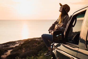 Seitenansicht einer Frau, die die Lichter des Sonnenuntergangs genießt und Gitarre spielt, während sie auf einem Auto am Meer sitzt. - ADSF06510