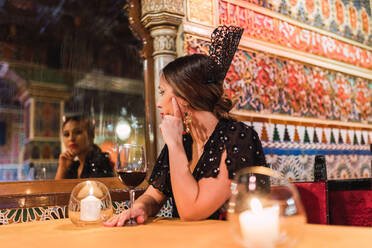 Attractive young lady in dress sitting at table near glass of drink and burning candles in luxury room of cafe decorated by mosaic - ADSF06520