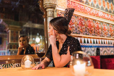 Attraktive junge Frau im Kleid sitzt am Tisch in der Nähe von Glas Getränk und brennende Kerzen in Luxus-Saal des Cafés mit Mosaik dekoriert - ADSF06520
