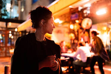 Trendiger junger Hipster mit Ohrringen, der in der Nähe eines Straßencafés bei Nacht in Tel Aviv, Israel, wegschaut - ADSF06527