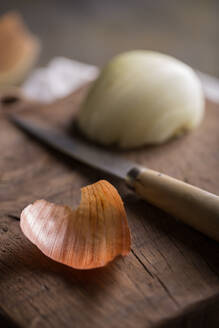 Nahaufnahme einer halb geschälten Zwiebel mit Schale und Küchenmesser auf einem Holzschneidebrett, symbolisiert die Zubereitung von Speisen und Kochkonzepte - ADSF06550