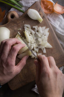 Nahaufnahme von Händen, die weiße Zwiebeln auf einem hölzernen Schneidebrett in der Küche hacken, mit grünen Zwiebeln im Hintergrund, die auf die Zubereitung einer Mahlzeit oder das Kochen zu Hause hinweisen. - ADSF06551