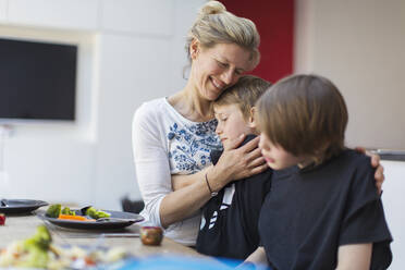 Affectionate mother hugging sons at dinner table - CAIF28827