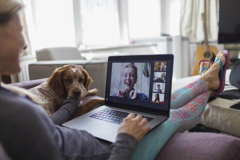 Woman with laptop video chatting with friends on sofa with dog - CAIF28843