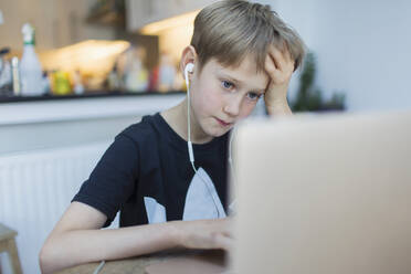 Focused boy with headphones using laptop in kitchen - CAIF28849