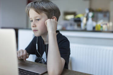 Konzentrierter Junge mit Kopfhörern bei der Hausaufgabenbetreuung am Laptop - CAIF28854