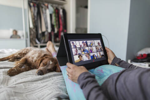 Frau mit digitalem Tablet im Videochat mit Freunden auf dem Bett mit Hund, lizenzfreies Stockfoto