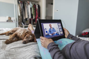 Woman with digital tablet video chatting with doctor on bed with dog - CAIF28889