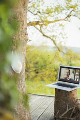 Gesichtsmaske hängt an einem Baum in der Nähe von Freunden, die auf einem Laptop-Bildschirm videochatten - CAIF28924