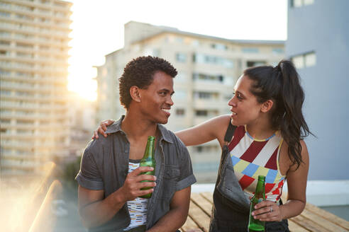Glückliches junges Paar trinkt Bier auf einem sonnigen städtischen Dachbalkon - CAIF29084