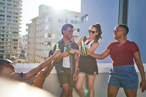 Glückliche junge Freunde stoßen mit Bierflaschen auf einem sonnigen städtischen Balkon an - CAIF29095