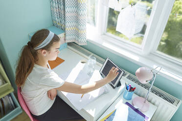 Mädchen mit digitalem Tablet bei der Hausaufgabenbetreuung am Schreibtisch im sonnigen Schlafzimmer - CAIF29114