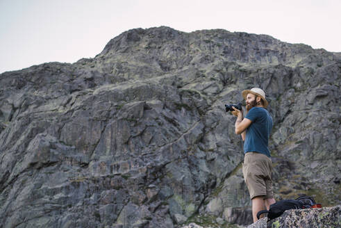 Seitenansicht eines jungen, gut aussehenden Mannes in Hut und T-Shirt, der in der Nähe einer hohen Felsklippe steht und ein Foto mit einer Fotokamera macht - ADSF06581