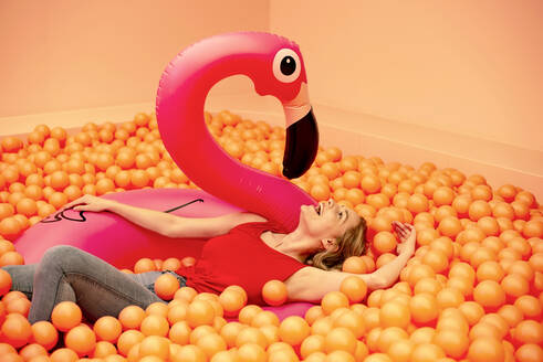 Cheerful young woman with inflatable flamingo lying in orange ball pit - BFRF02272