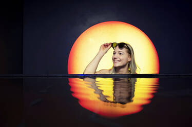 Lächelnde junge Frau mit Sonnenbrille schwimmt im Pool vor dem Hintergrund des Sonnenuntergangs - BFRF02274