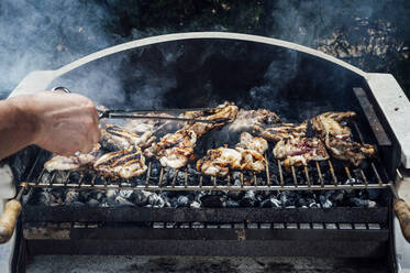 Hand of young man cooking meat on barbecue grill in yard - JCMF01052