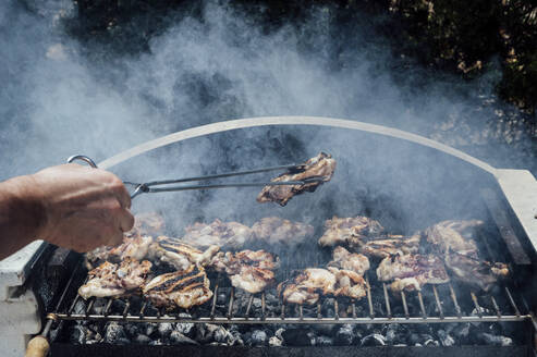 Die Hand eines jungen Mannes hält Fleisch mit einer Zange auf einem Grill im Hof - JCMF01053