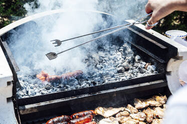 Die Hand eines jungen Mannes hält eine Servierzange über einem Grill im Hof - JCMF01059