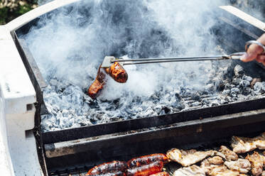 Die Hand eines jungen Mannes hält Fleisch mit einer Servierzange über einem Barbecue-Grill im Hof - JCMF01060