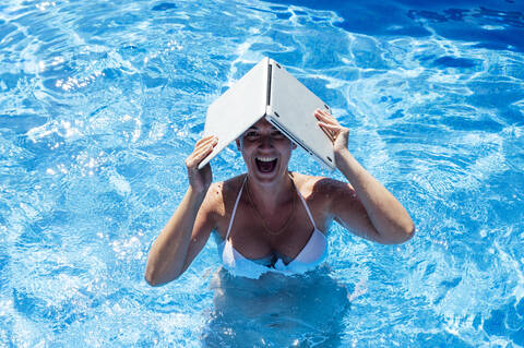 Cheerful woman holding laptop on head while swimming in pool stock photo