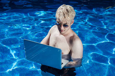 Shirtless young man wearing sunglasses using laptop while standing in swimming pool - JCMF01068