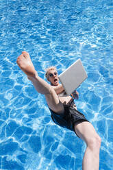 Shirtless young man holding laptop falling in swimming pool - JCMF01069